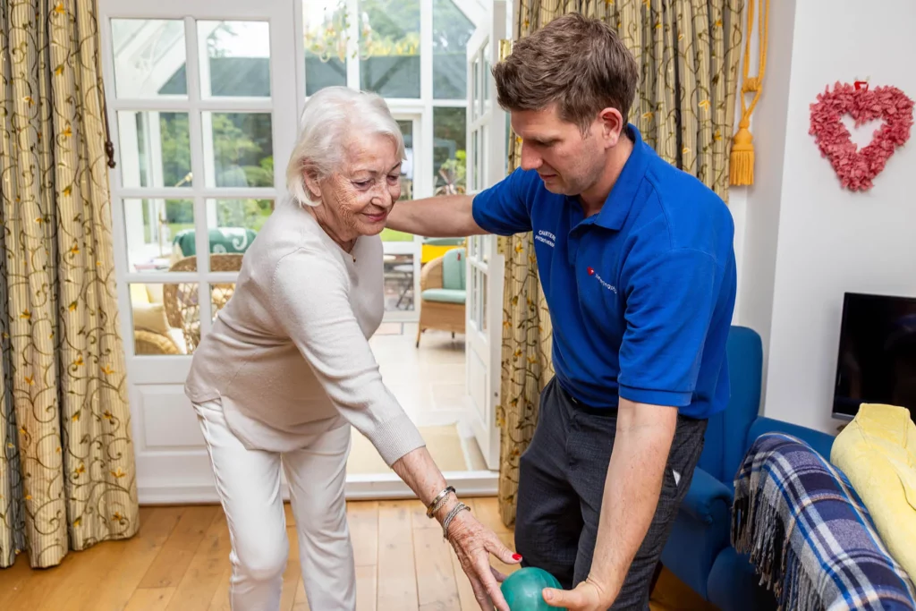 Physiotherapist supporting an older woman as she walks with a frame in her conservatory at home