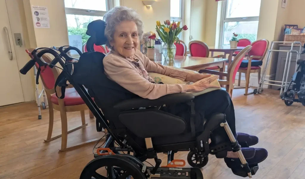 An older woman with dementia sitting comfortably in a specialist postural wheelchair in a care home lounge