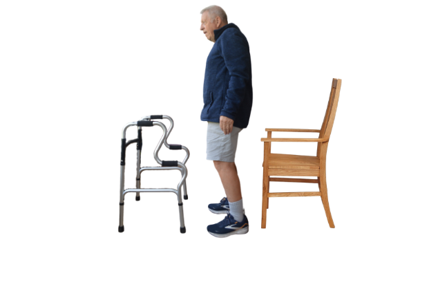 Older man standing upright with a walking frame in front of him and a chair directly behind