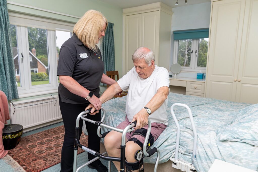 A physiotherapist steadying a walking frame as an older man stands up from the side of his bed