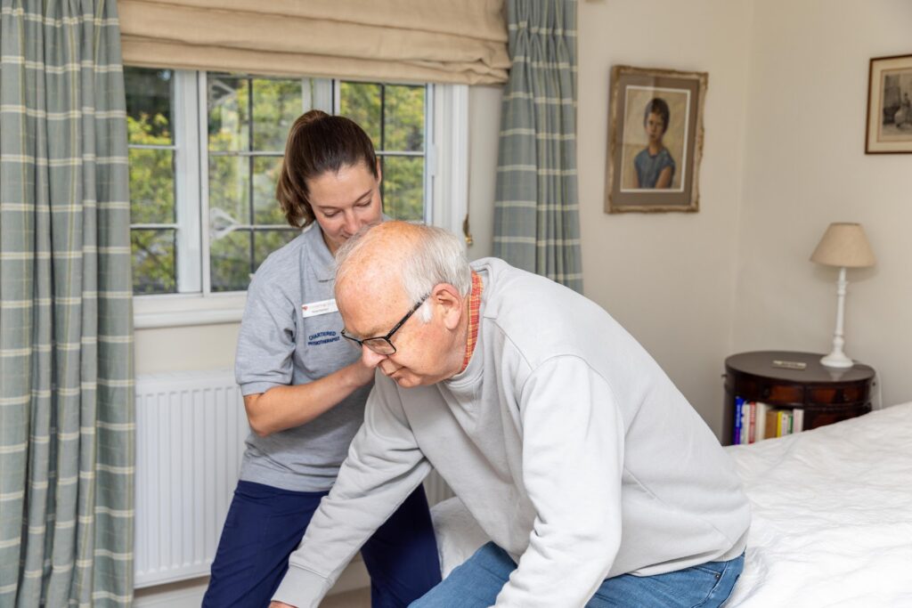 A physiotherapist supporting an older man as he leans forward to get up from his bed