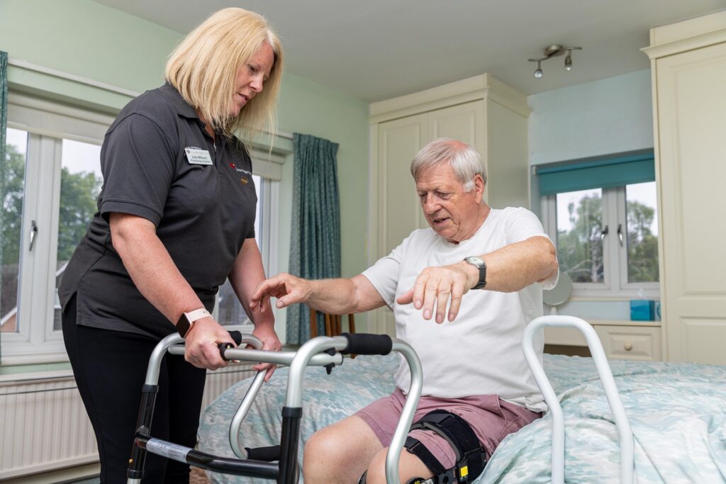 A physiotherapist talking with an older man as he stands with a walking frame in his bedroom