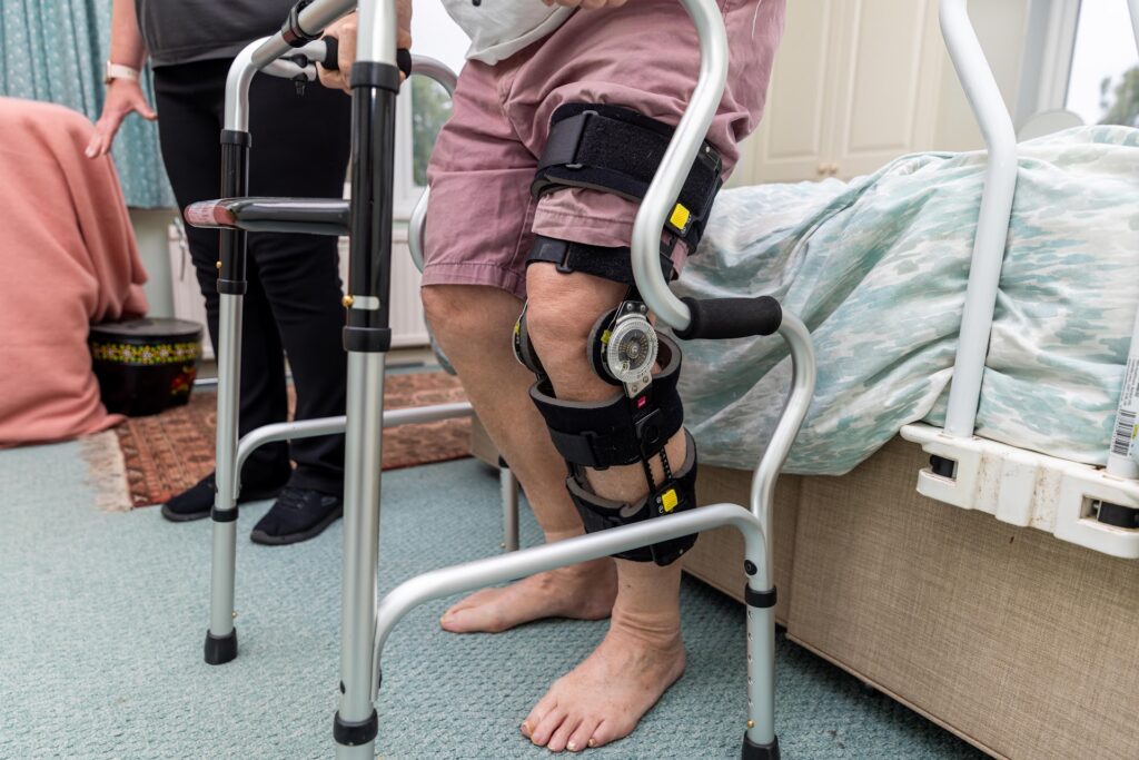 An older man wearing a hinged knee brace standing at his bedside with a walking frame, supported by a physiotherapist