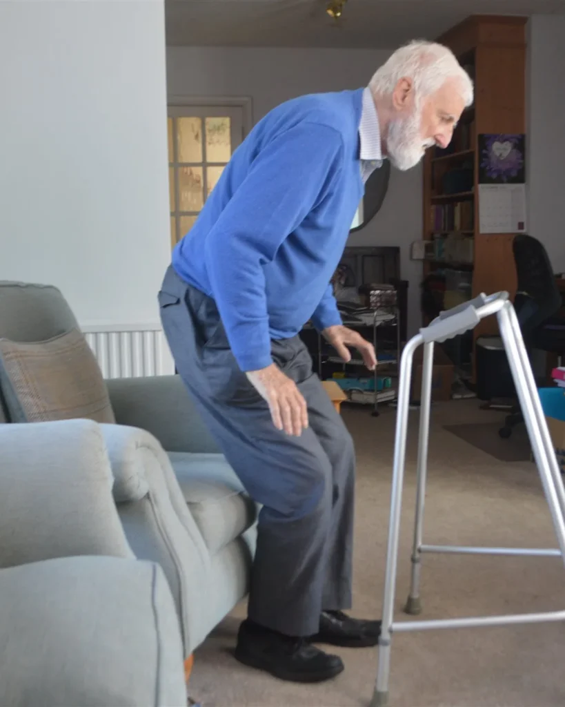 Older man rising from an armchair and reaching for a walking frame