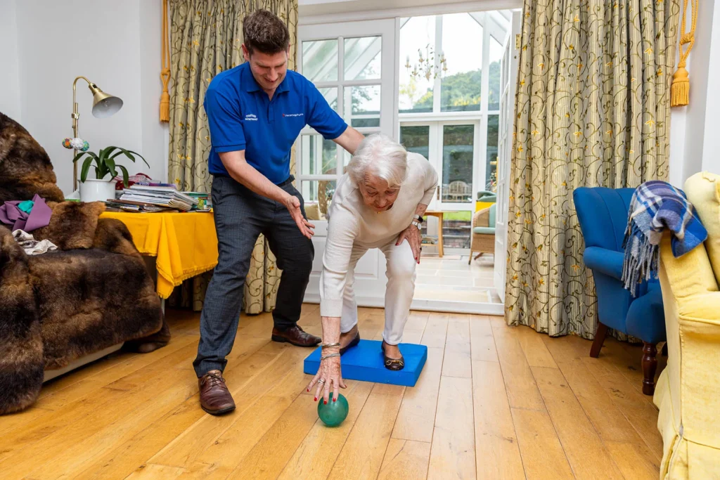 A physiotherapist steadying an older woman as she bends forward from a balance pad to pick up a ball, during a home visit physiotherapy session in Woking.